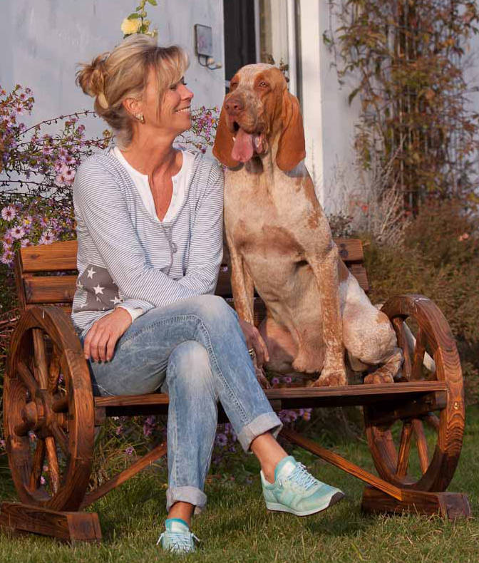 Susa Gülzow sitzt lächelnd auf einer Holzbank im Garten, neben ihr ein orange-weißer Jagdhund